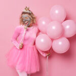 carefree-cheerful-young-woman-with-beauty-clay-mask-on-face-holds-magic-wand-near-mouth-as-if-microphone-wears-dress-celebrates-festive-event-holds-bunch-of-inflated-balloons-holiday-concept